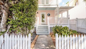 A white picket fence, a concrete pathway, and a pink building with a white porch.