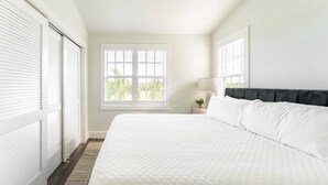 A bedroom with a large bed, a window with blinds, a bedside table with a lamp, and a dark headboard.