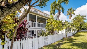 A two-story house with a white picket fence, surrounded by lush greenery and palm trees.