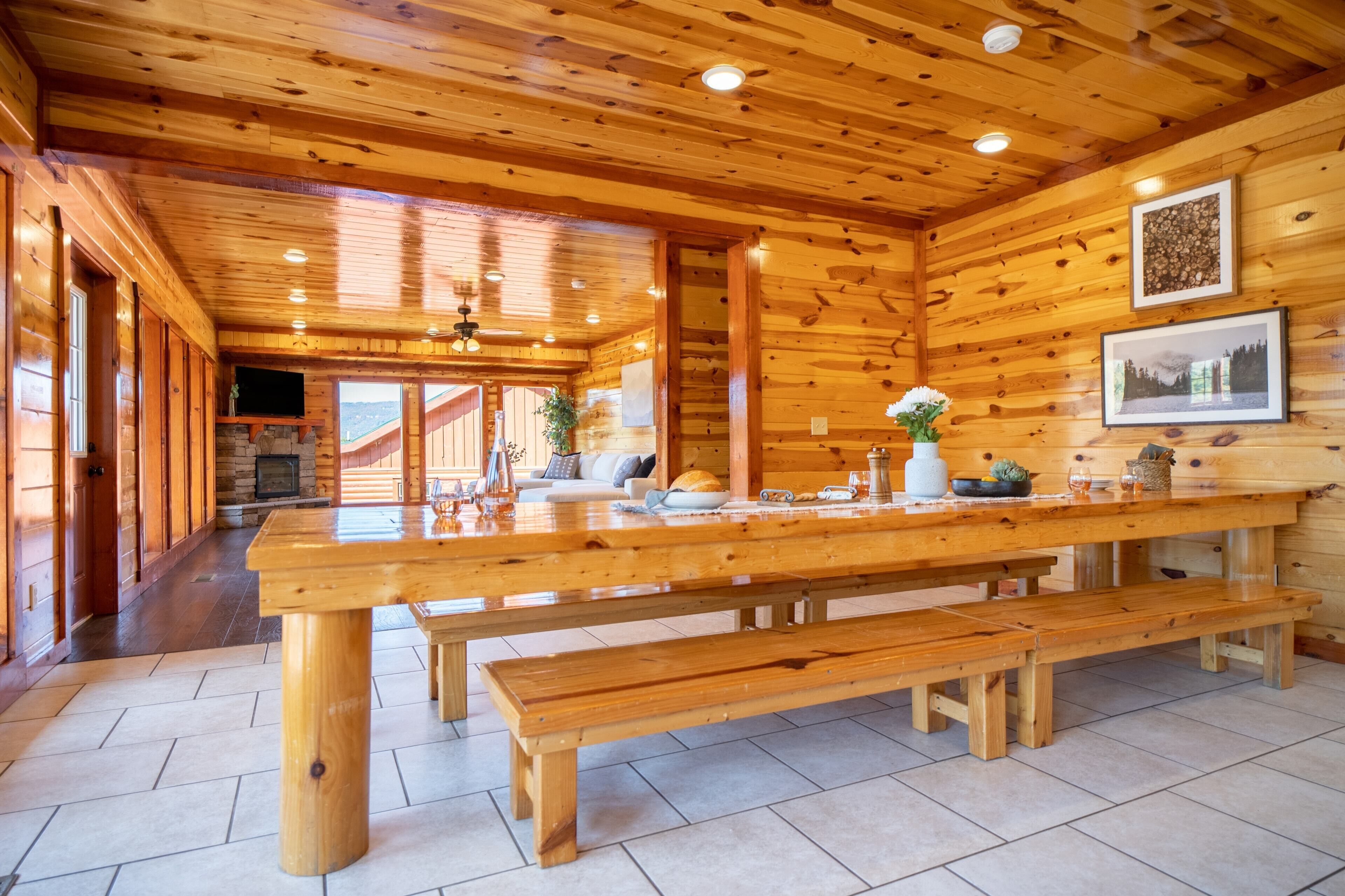A spacious wooden cabin interior with a large dining table, benches, a stone fireplace, and framed pictures on the walls.