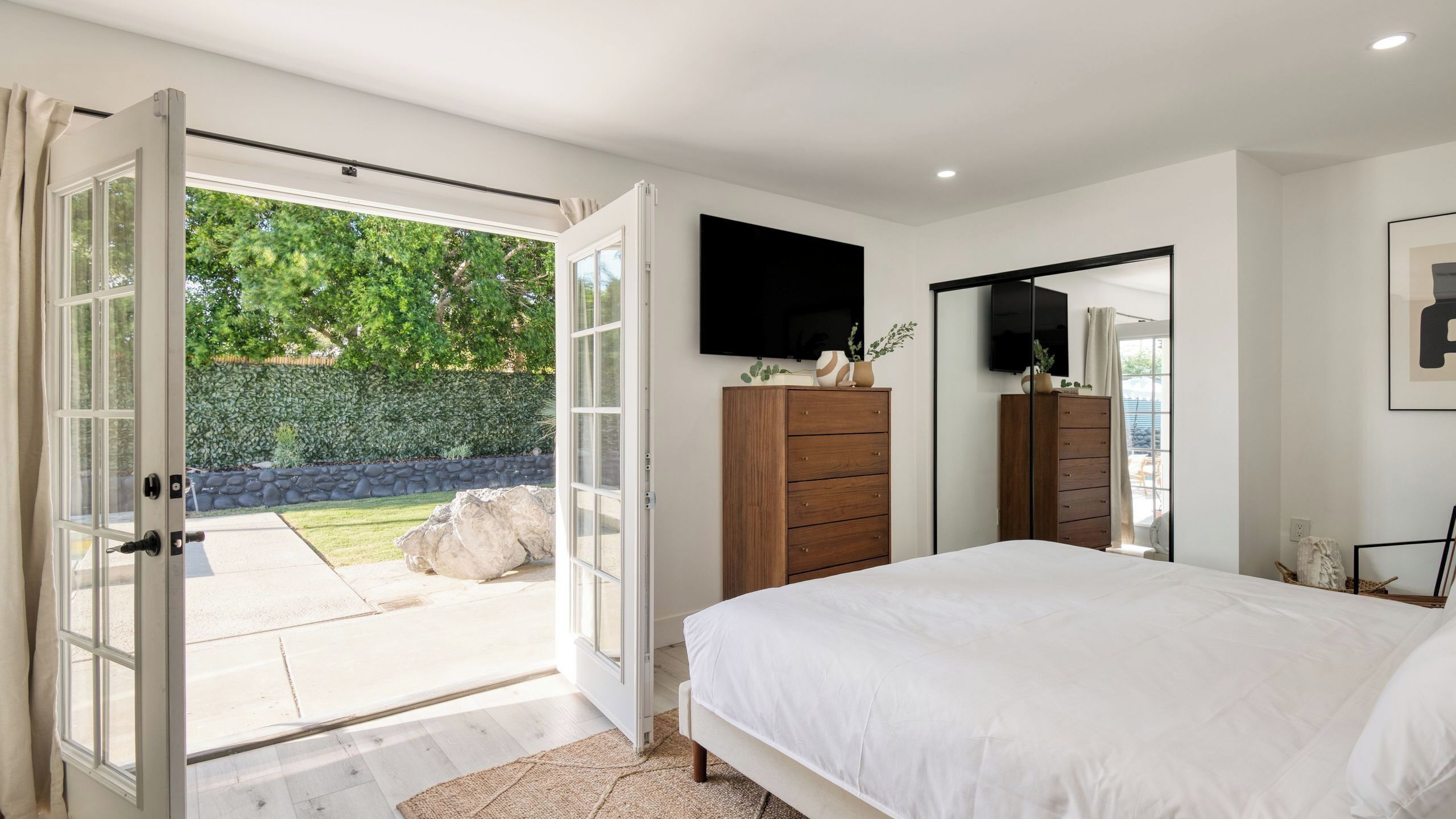 A bedroom with a large bed, a TV mounted on the wall, and a sliding door leading to a garden.