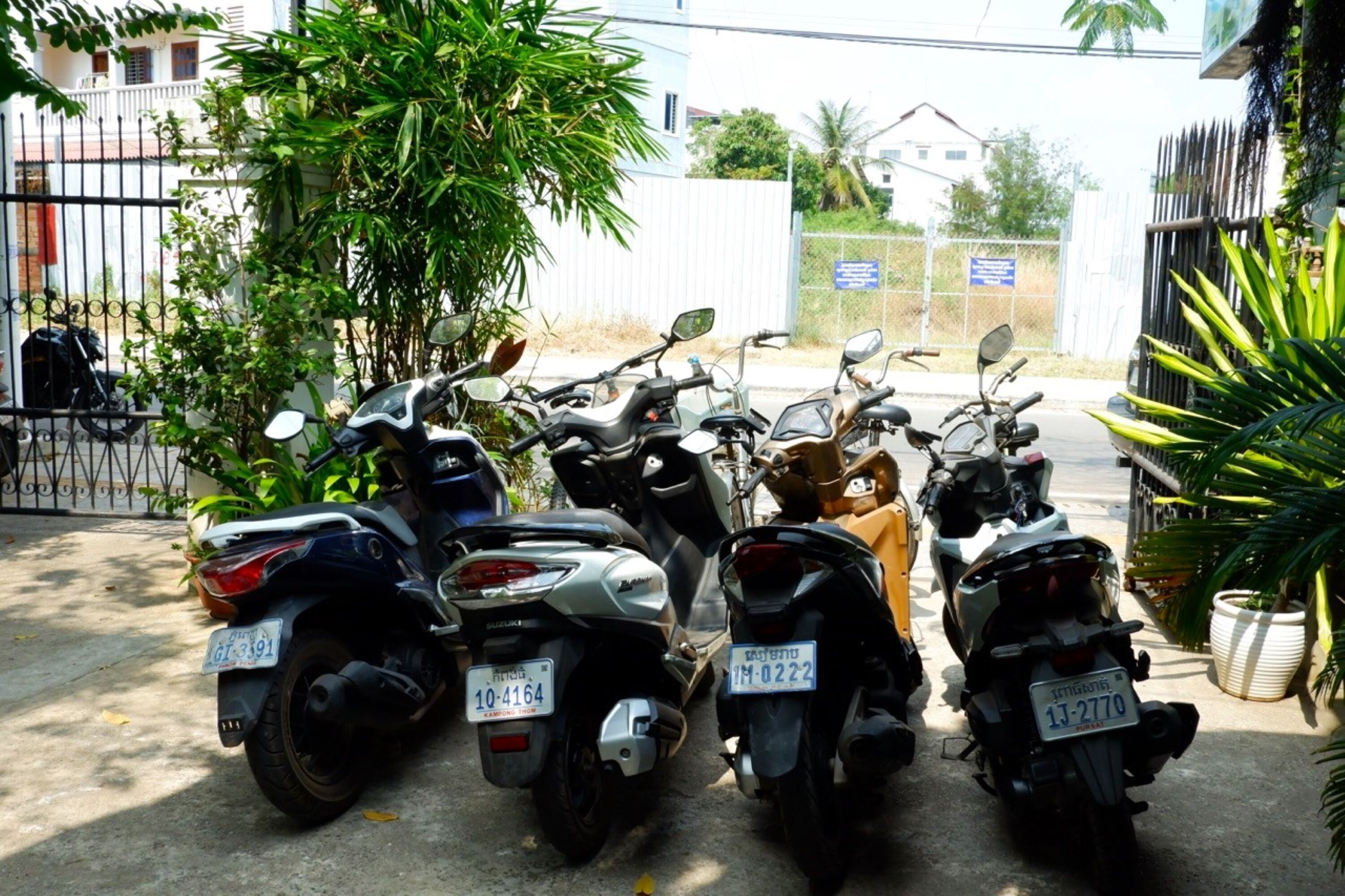 A row of parked motorcycles in an outdoor area with a white fence and greenery.