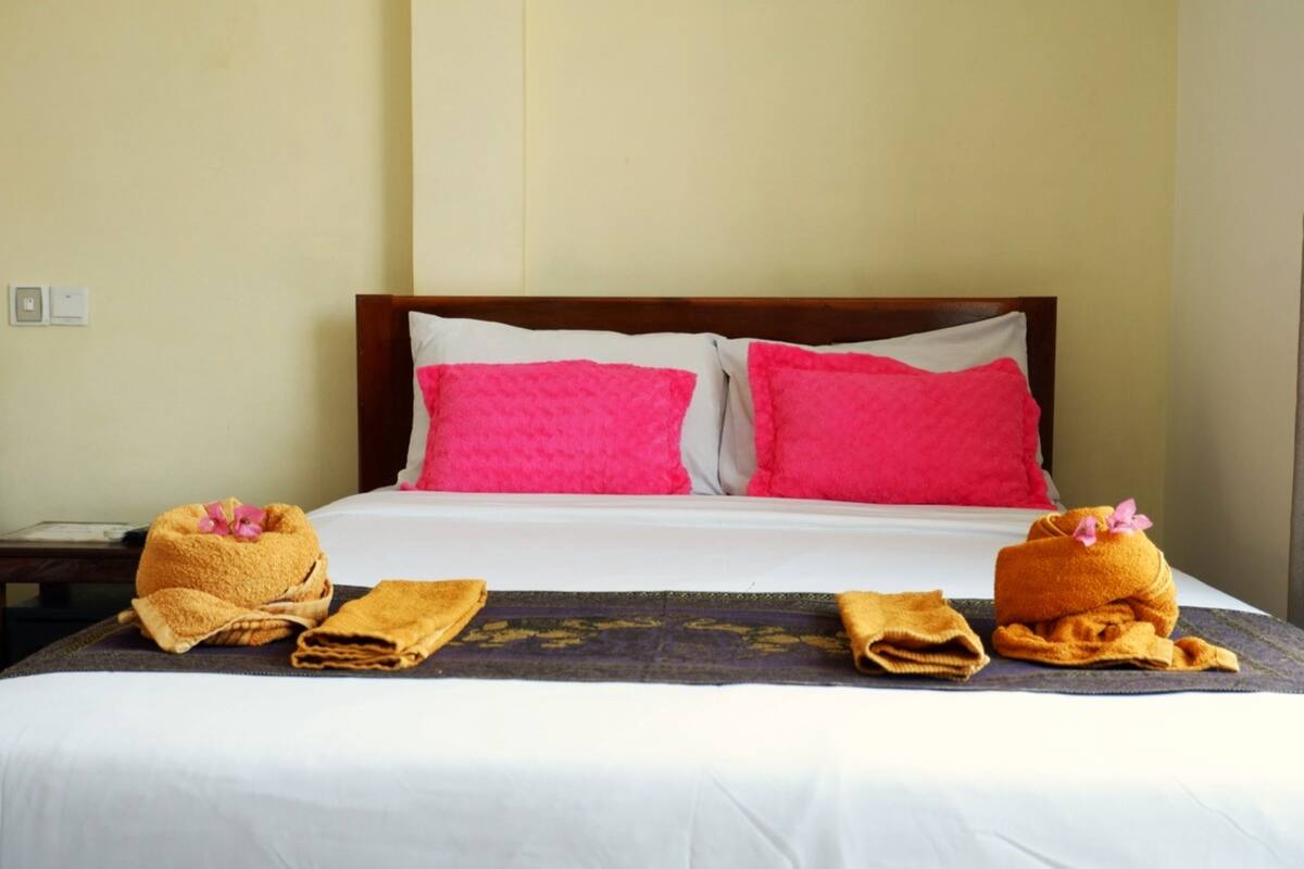 Ein Hotelzimmer mit einem ordentlich bezogenen Bett, zwei hellrosa Kissen und zwei Handtüchern mit einer rosa Blume darauf.