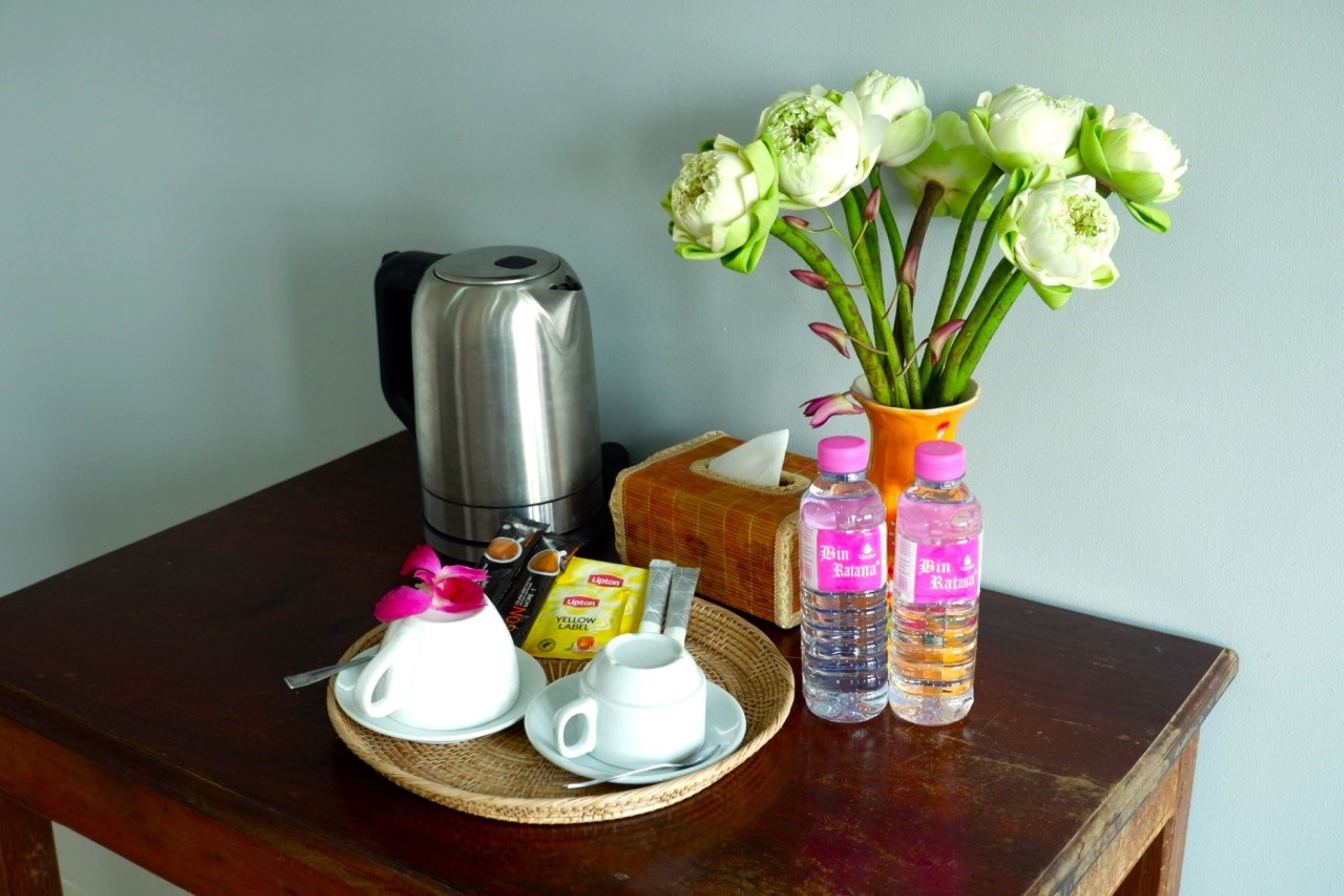 A breakfast setup with a coffee maker, a vase of flowers, a tissue box, a water bottle, and a packet of instant coffee.