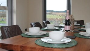 A dining table set with white bowls and plates, a bottle of champagne, and glasses, with a view of a vineyard through large windows. - Forest View House - Private Hot Tub (Coleraine)