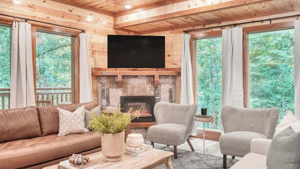 L’intérieur chaleureux d’un chalet avec une cheminée en pierre, un téléviseur à écran plat, un canapé en cuir marron et deux fauteuils gris.
