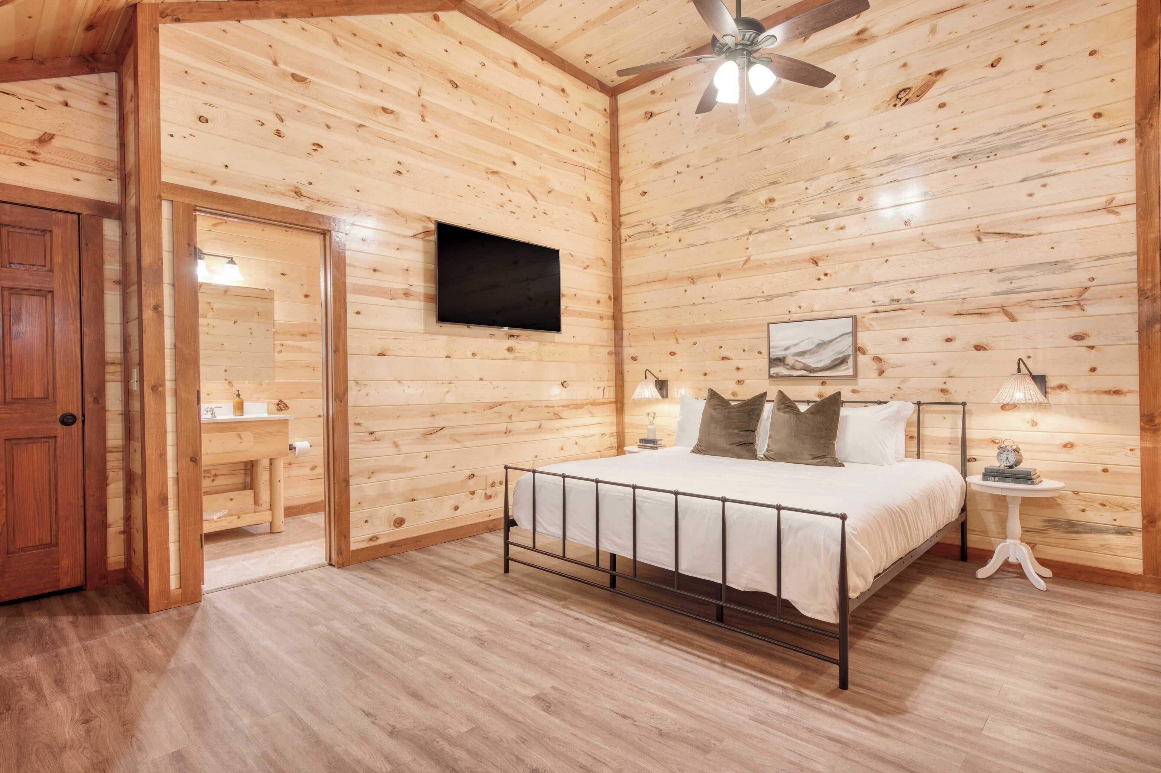 Une chambre à coucher aux murs lambrissés, avec un lit, un ventilateur de plafond, un téléviseur fixé au mur et une table de chevet avec une lampe.