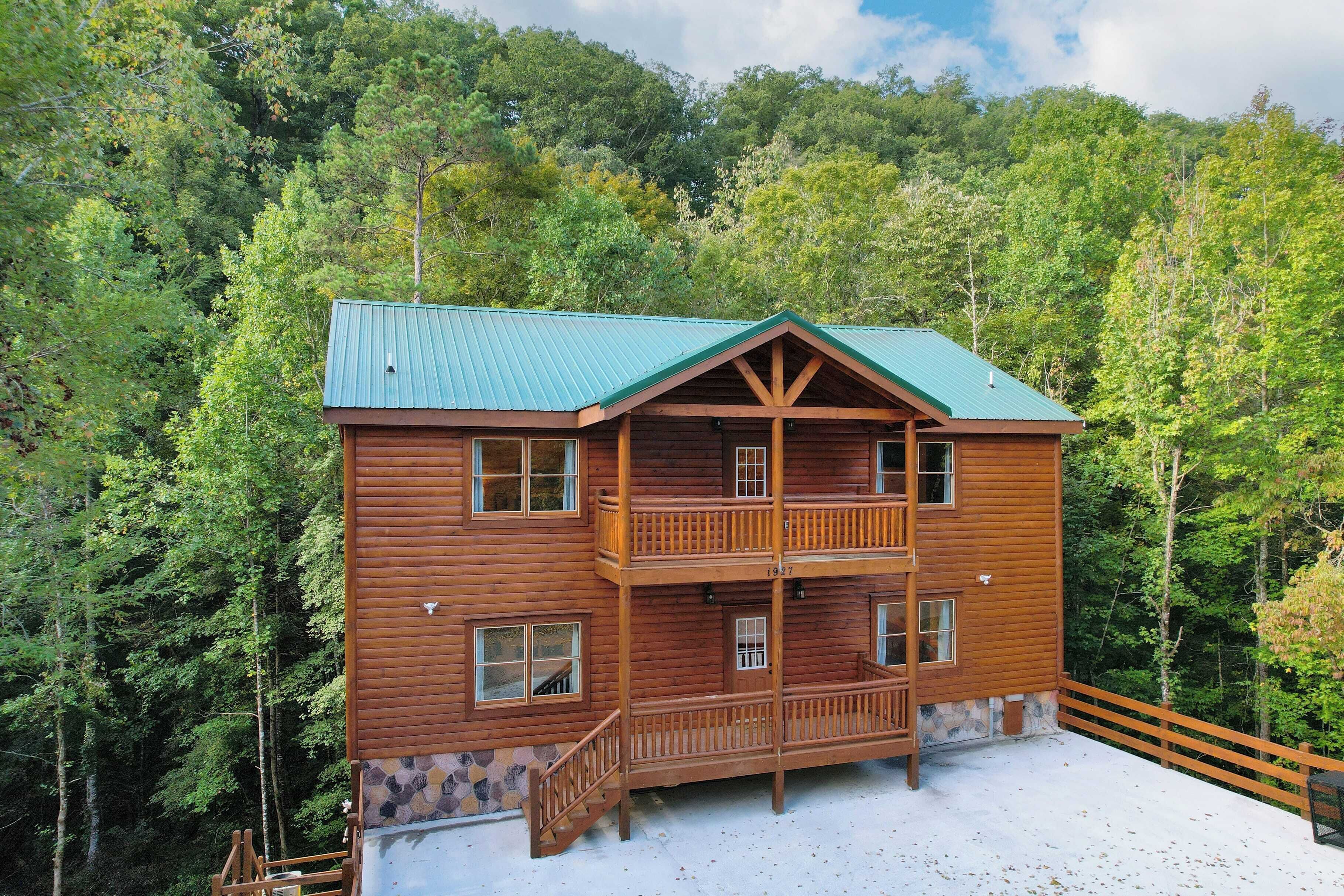 A two-story log cabin with a green roof, surrounded by dense forest.