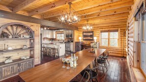 A log cabin interior with a wooden dining table, chairs, a kitchen area, and a chandelier.
