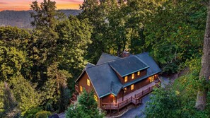 A two-story log cabin with a balcony, surrounded by dense forest. - Leyland by Avantstay Cabin Mansion w/ Views (Asheville)