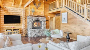 A cozy log cabin interior with a stone fireplace, wooden walls, a staircase, and a comfortable seating area.