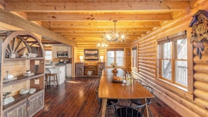 A log cabin interior with a wooden dining table, kitchen area, and large windows.