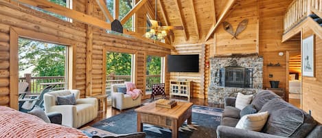 A log cabin interior with a fireplace, wooden furniture, and large windows.