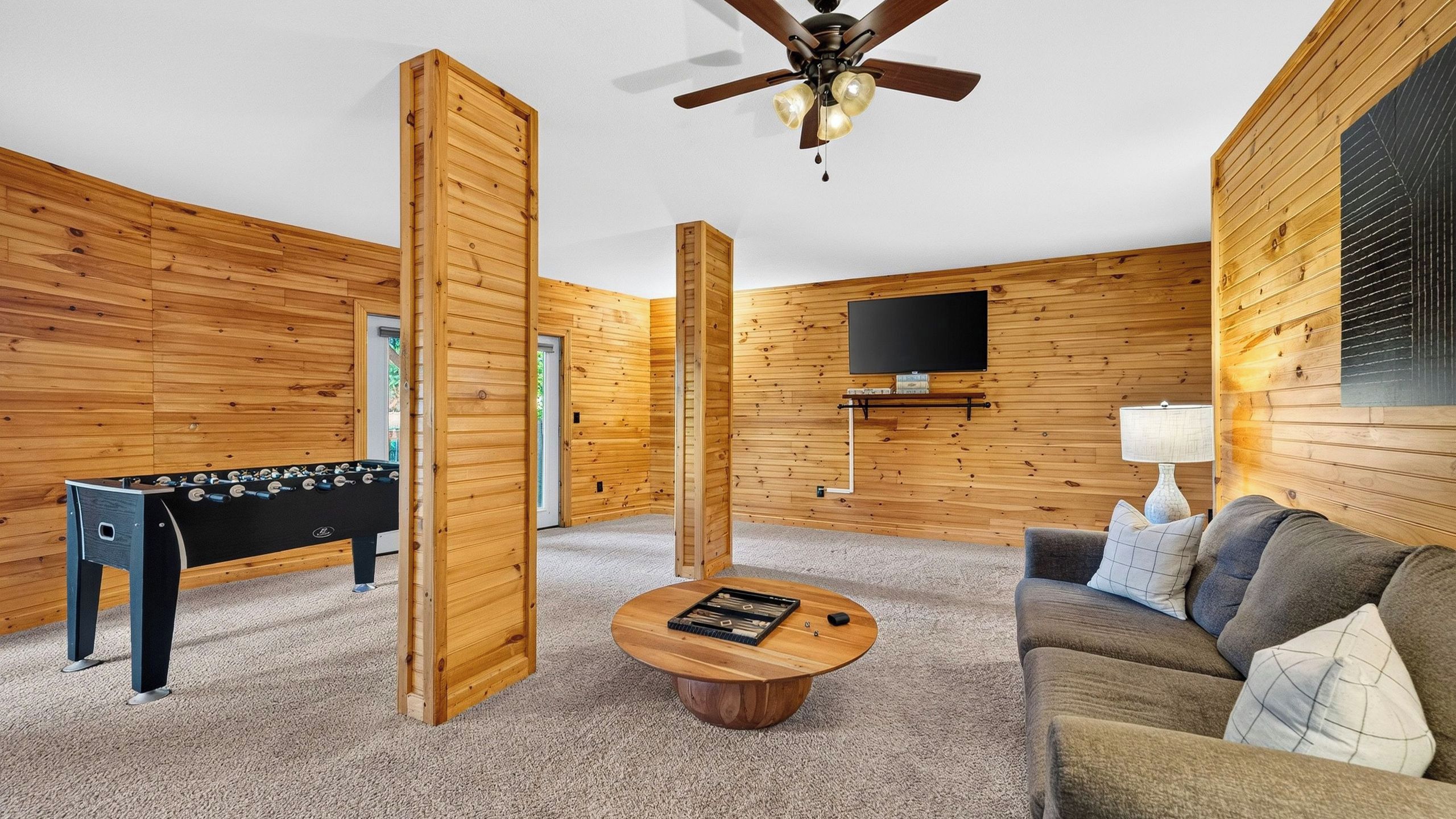 A room with wooden walls, a ceiling fan, a foosball table, a coffee table, and a sofa with cushions.
