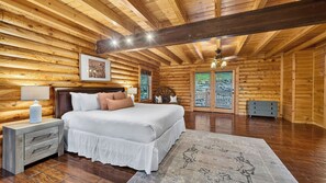 A log cabin bedroom with a bed, bedside table, lamp, and a view through the window. - Leyland by Avantstay Cabin Mansion w/ Views (Asheville)