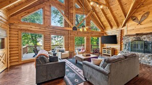A log cabin interior with a fireplace, wooden ceiling, and large windows. - Leyland by Avantstay Cabin Mansion w/ Views (Asheville)