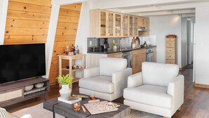 Ein modernes Wohnzimmer mit Fernseher, zwei Sesseln, einem Couchtisch und einer Küche im Hintergrund.