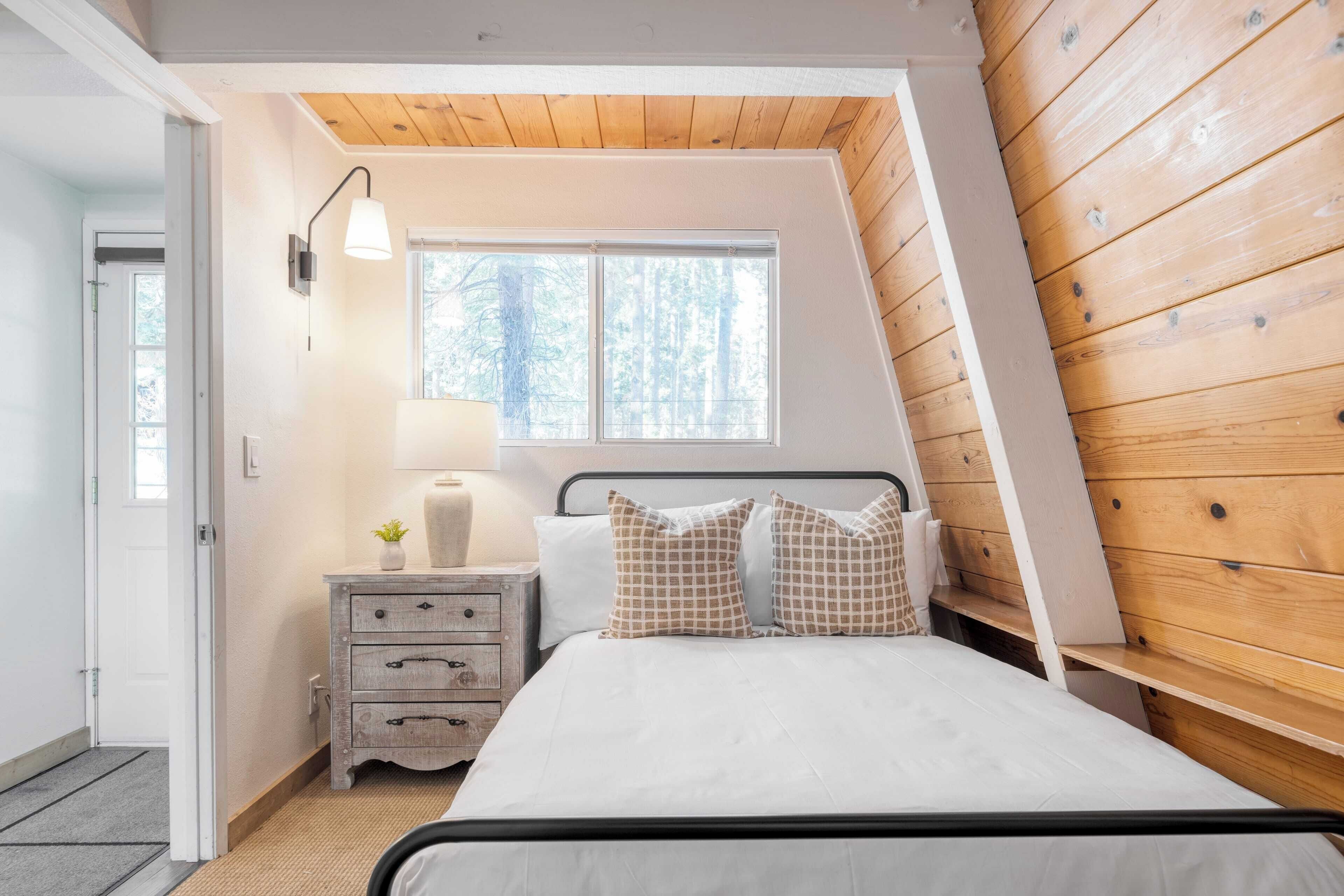 A bedroom with a sloped ceiling, a bed with white bedding, a bedside table with a lamp, and a window with a view of trees.