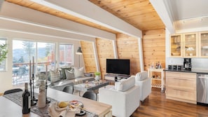 Amplia sala de estar con techo de madera, televisor de pantalla plana, mesa de comedor con aperitivos y cocina con armarios de madera y máquina de café.