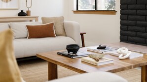 A cozy living room with a sofa, a wooden coffee table with books and decorative items, a fireplace, and a window with a view of greenery. - Archie by Avantstay Desert Retreat w/ Pool, Edge of Joshua Tree, Bonus Casita for Group Travel (Yucca Valley)