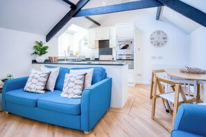 A modern kitchen with white cabinets, a built-in oven, and a dining area with a wooden table and chairs. - Penthouse At The Mews - 2 Bed Apartment - Tenby (Tenby)