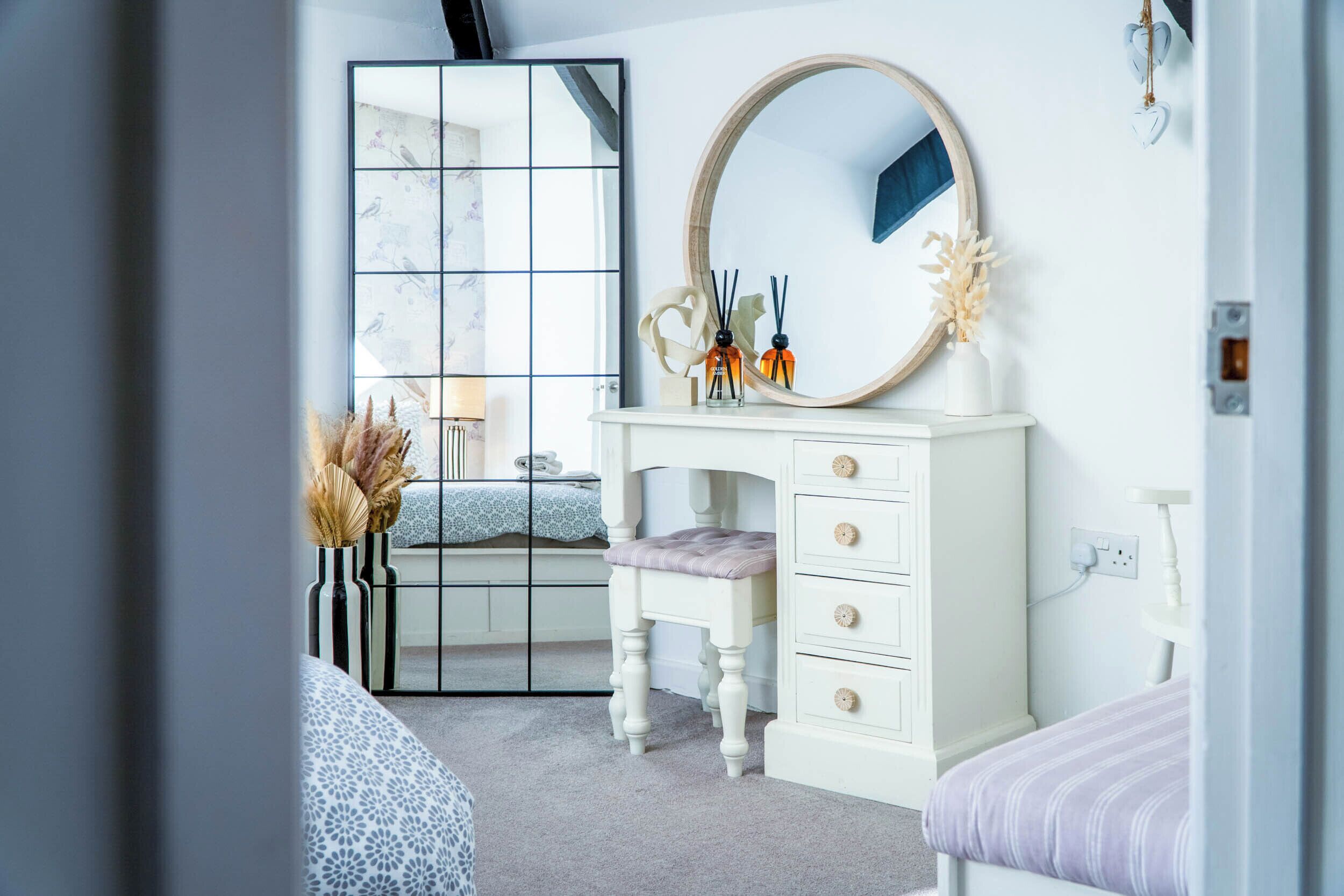 Ein Schlafzimmer mit weißem Schminktisch, einem runden Spiegel, einem Fenster mit Gittermuster und einem Bett mit blau-weiß gestreifter Bettdecke.