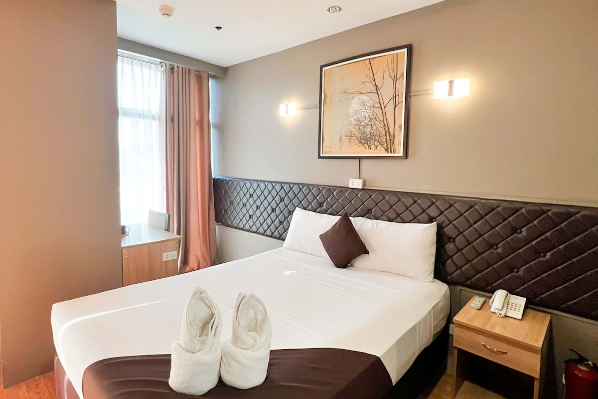 Une chambre d’hôtel avec un grand lit, une table de chevet avec un téléphone, une lampe fixée au mur et une fenêtre avec des rideaux.