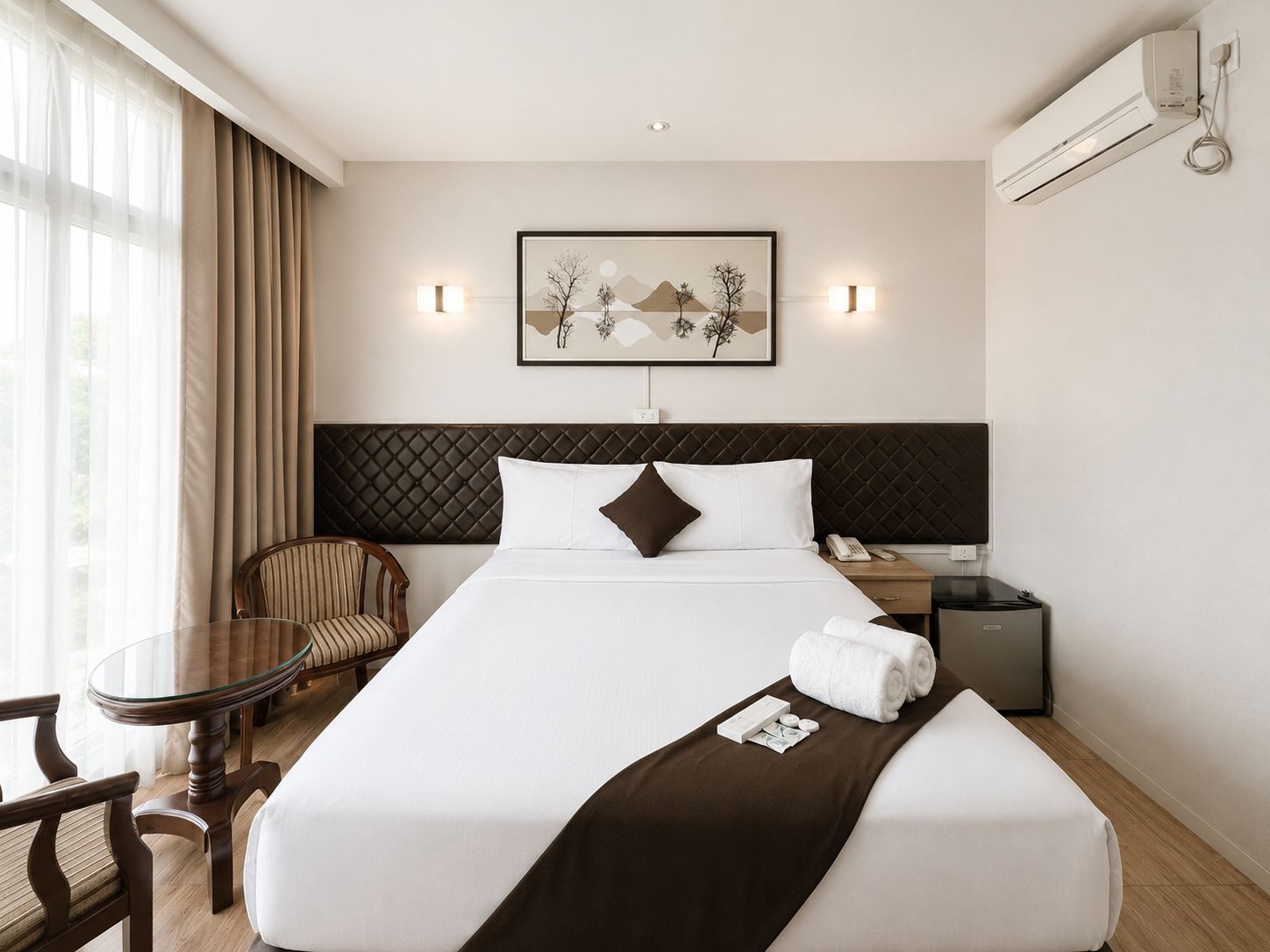 A hotel room with a bed, a chair, a table, a bedside table, a lamp, and a wall-mounted air conditioner.