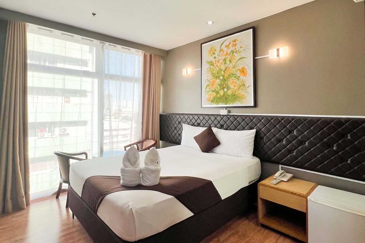 Une chambre d’hôtel avec un grand lit, une table de chevet avec un téléphone, une chaise et une fenêtre avec des rideaux.