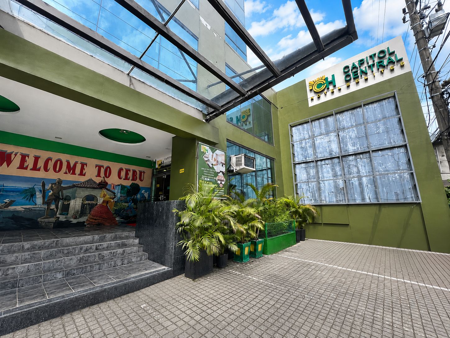 Entrada de un hotel con un mural que dice 'Welcome to Cebu', fachada de vidrio y un letrero que indica 'Capitol Central Hotel'.