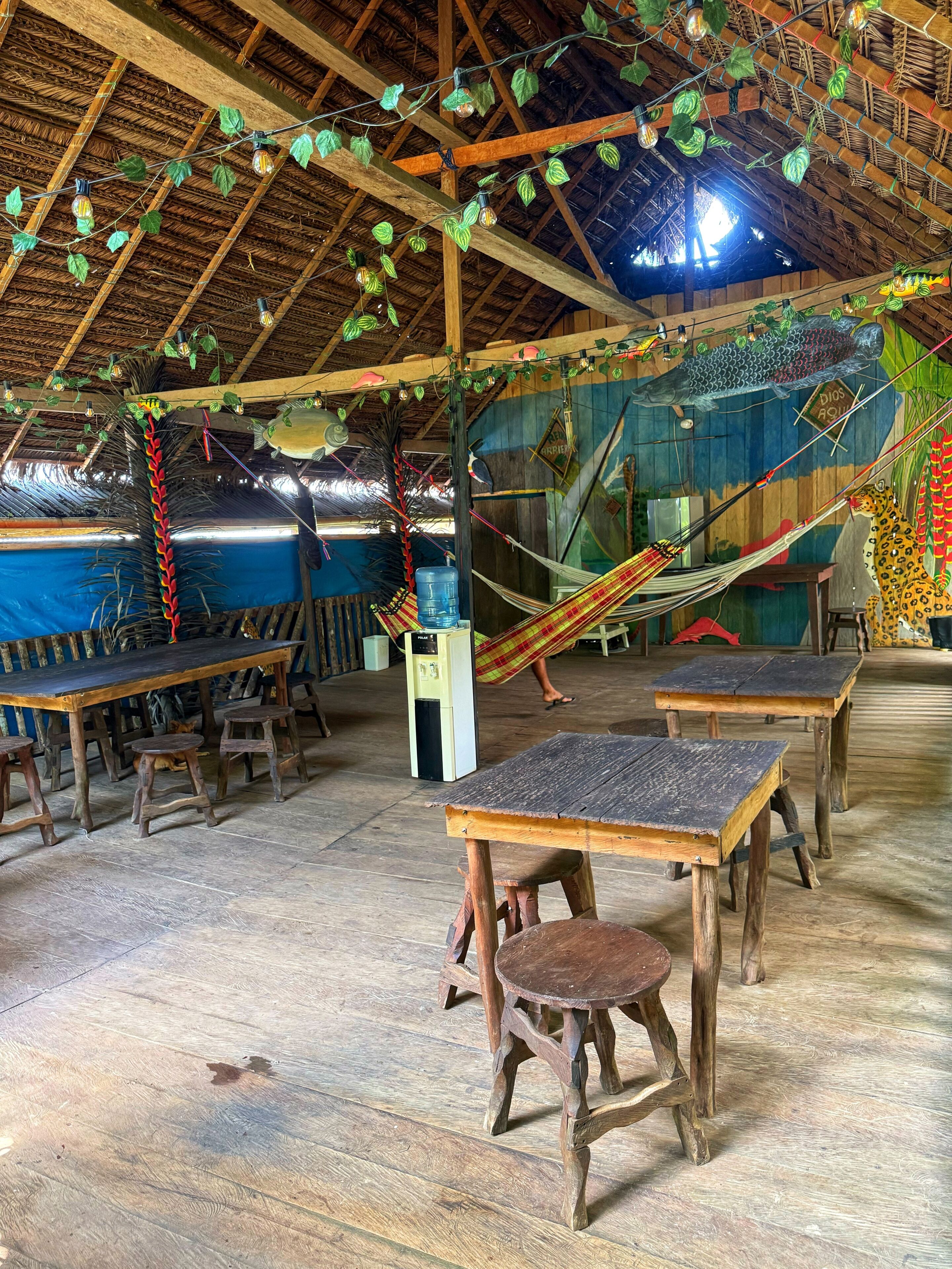 An indoor space with wooden tables and benches, a hammock, and a thatched roof ceiling.