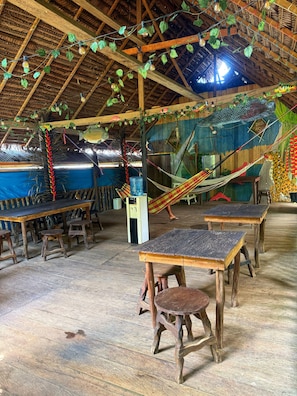 Ein Innenraum mit Holztischen und -bänken, einer Hängematte und einer Strohdachdecke.
