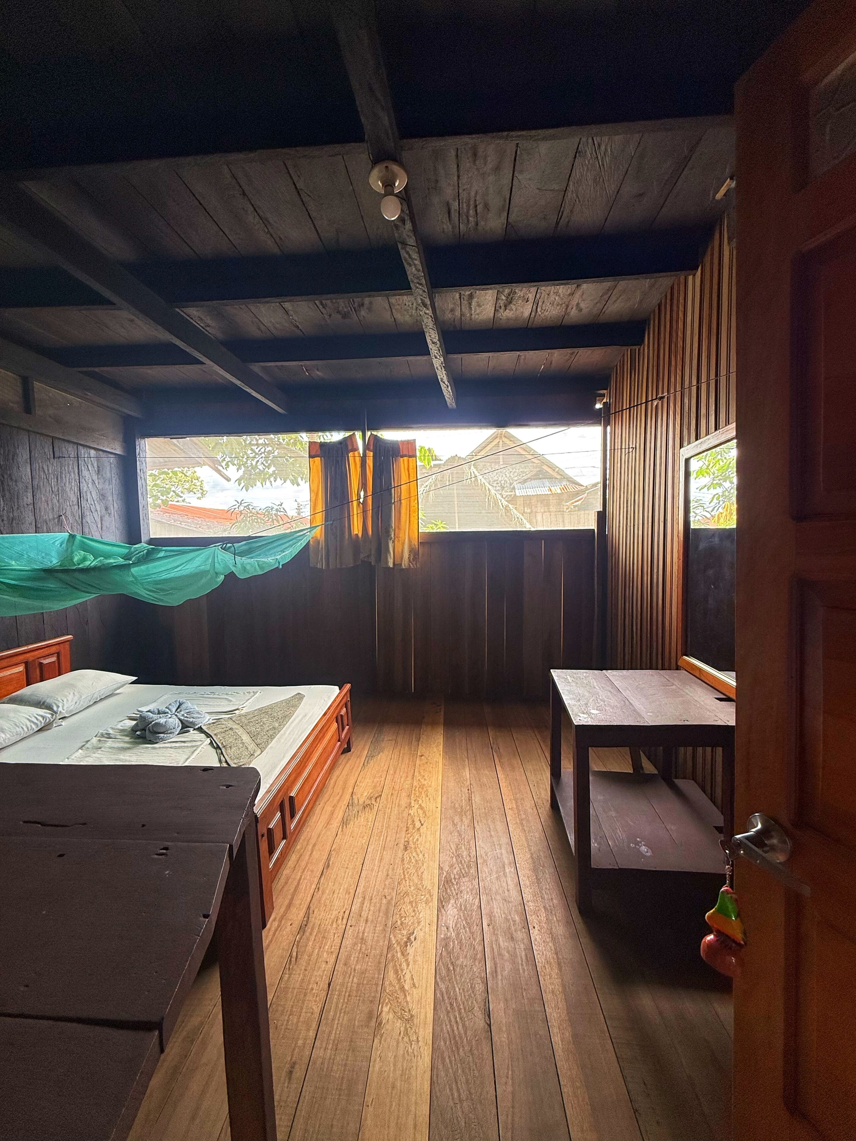 A room with a wooden ceiling, a bed with a wooden frame, a table, and a view of the outside.
