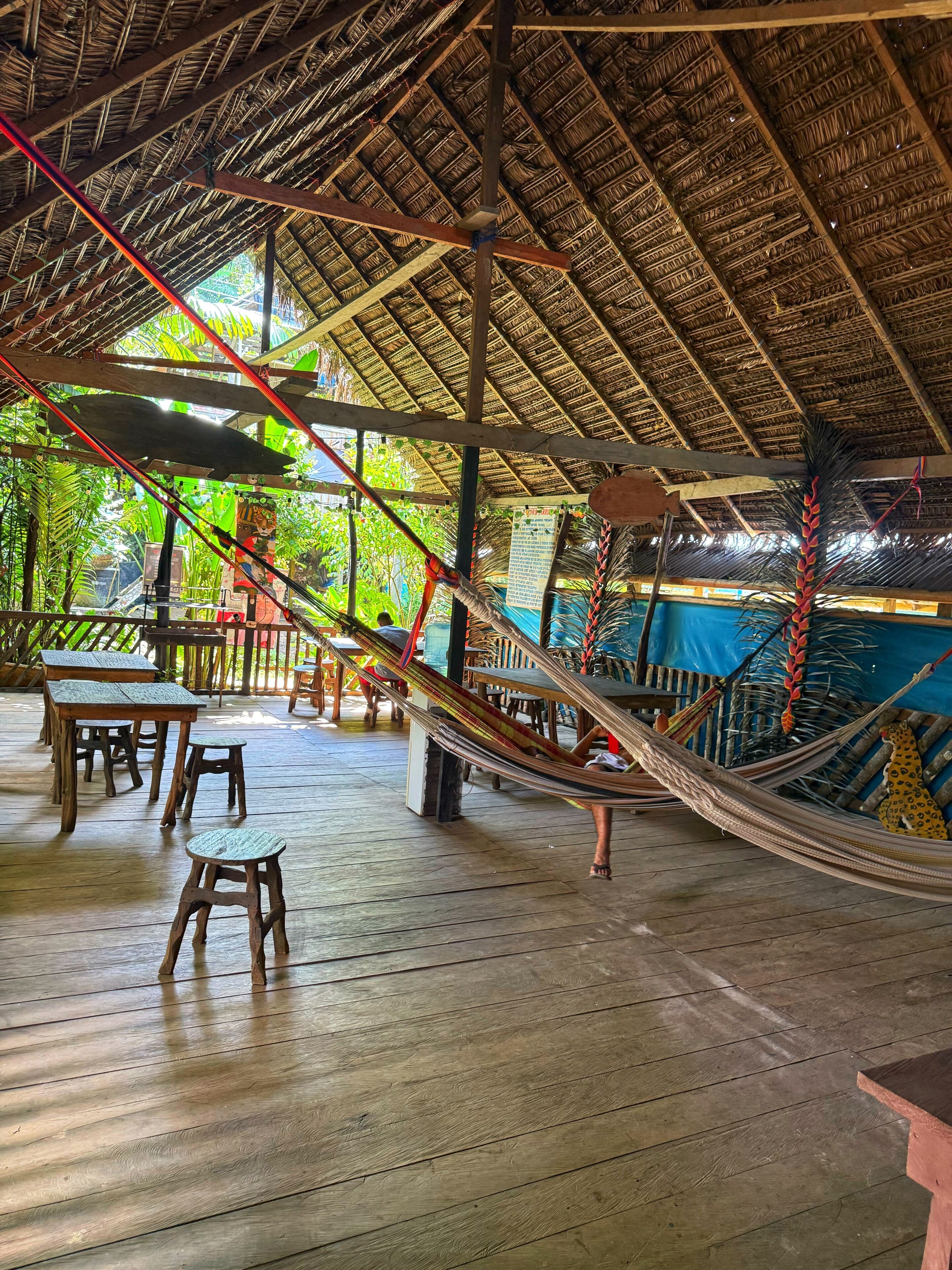 A covered outdoor area with wooden tables and chairs, a hammock, and a thatched roof structure.