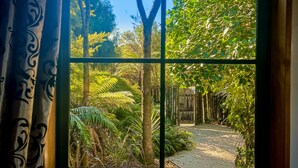 A view through a window with a patterned curtain, revealing a garden path surrounded by lush greenery and trees. - Te Tiro Accommodation (Waitomo Caves)