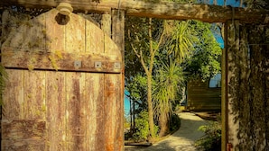 A rustic wooden gate with a circular window, leading to a pathway surrounded by lush greenery and a small building. - Te Tiro Accommodation (Waitomo Caves)