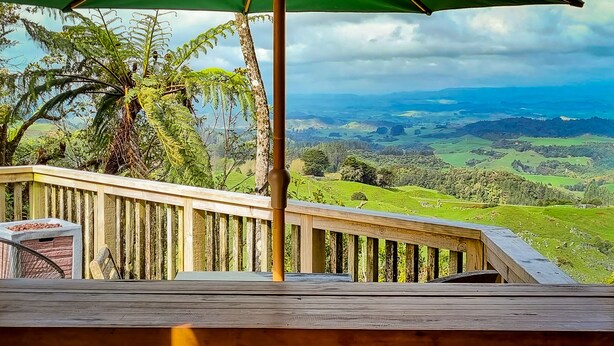 A wooden deck with a green umbrella, overlooking a lush green landscape with hills and trees. - Te Tiro Accommodation (Waitomo Caves)