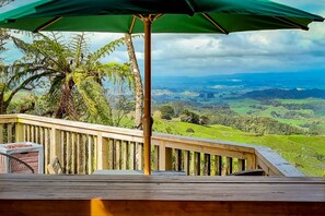 A wooden deck with a green umbrella, overlooking a lush green landscape with hills and trees. - Te Tiro Accommodation (Waitomo Caves)