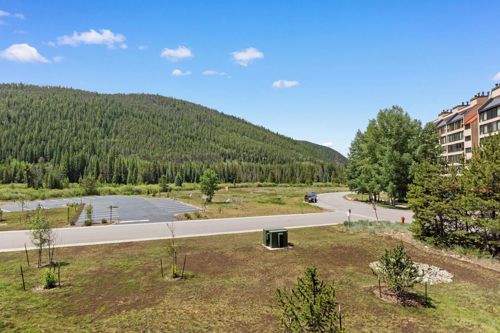 A parking lot with a road leading to a building surrounded by trees and a forested hill.