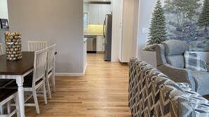 A dining area with a wooden table and chairs, a sofa with a patterned throw, and a hallway leading to a kitchen.