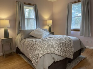 A bedroom with a bed, two bedside lamps, a window with curtains, and a wooden floor.