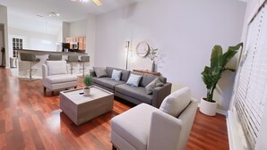Uma sala de estar moderna com sofá, poltronas e mesa de centro. Uma cozinha visível ao fundo, com banquetas de bar.