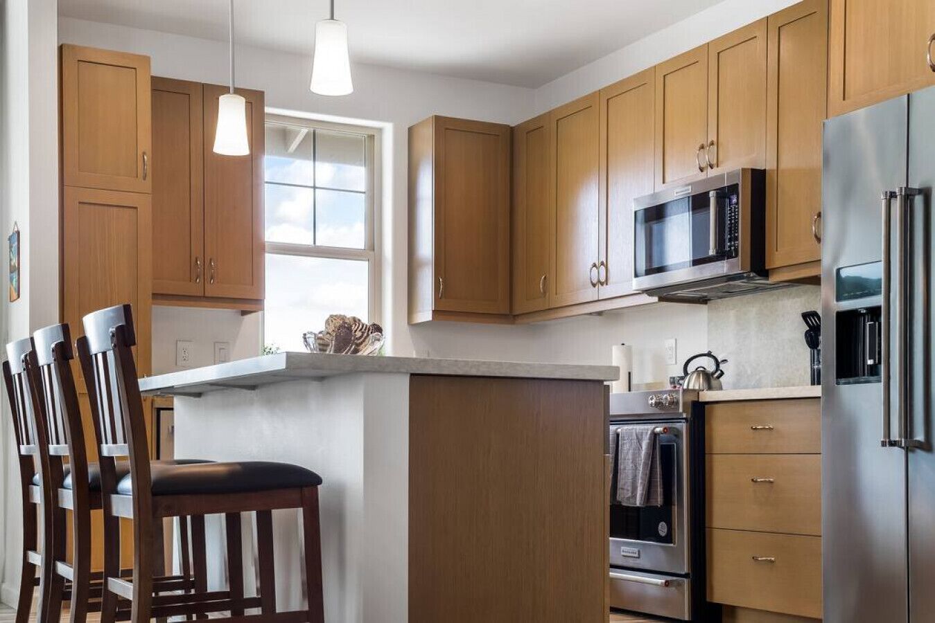 Una cocina moderna con armarios de madera, microondas integrado, refrigerador de acero inoxidable y un amplio ventanal que proporciona luz natural.