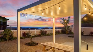 A covered outdoor patio with string lights, a wooden table, and a view of the ocean at sunset. - Mountain View|Single Lvl Home|Beach 5 Min|Free Prk (Waianae)