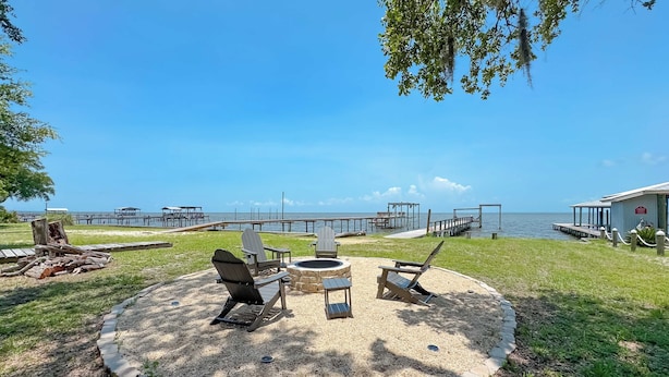 A circular seating area with a fire pit, surrounded by Adirondack chairs, overlooking a lake with docks and a wooden structure.