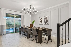 A dining area with a large table set for six, a modern chandelier, and a view of a swimming pool through glass doors. - Lakeview 9BR | Spider-Man Game Room, Pool & Spa (Kissimmee)