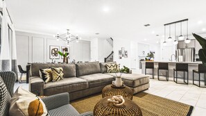 A modern living room with a grey sofa, patterned cushions, and a woven ottoman. - Lakeview 9BR | Spider-Man Game Room, Pool & Spa (Kissimmee)