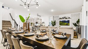 A modern dining area with a long wooden table set for six, surrounded by chairs. A kitchen and living area are visible in the background. - Lakeview 9BR | Spider-Man Game Room, Pool & Spa (Kissimmee)