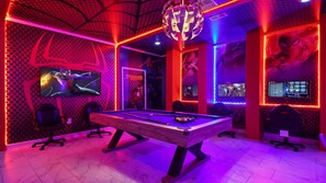 Una mesa de billar en una sala con paredes de diseño hexagonal, varias pantallas de juego y una lámpara de araña.