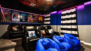 A home theater room with a row of black leather recliners, a blue bean bag chair, and a wall decorated with movie posters. - Lakeview 9BR | Spider-Man Game Room, Pool & Spa (Kissimmee)